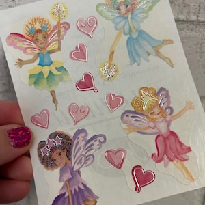 May include: Sheet of fairy stickers with watercolor illustrations. The fairies have colorful wings and dresses in shades of pink, blue, and purple. Pink heart stickers are also included.
