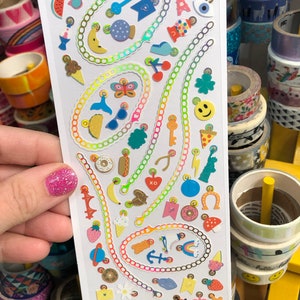 May include: A sheet of colorful holographic stickers with various designs, including a watermelon slice, a key, a butterfly, a hot dog, a donut, an anchor, a rainbow, and a ladybug. The stickers are arranged in a circular pattern around a chain link design. The text "YOU CHARMED ME" and "www.PIPSTICKS.com" is printed at the bottom of the sheet.