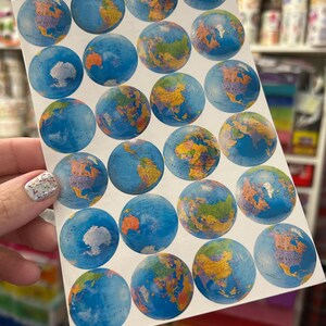 May include: A white sticker sheet featuring twenty-two circular globe decals. Each sticker displays a detailed map of the Earth with blue oceans and multicolored continents. The globes are arranged in a grid pattern across the rectangular sheet.