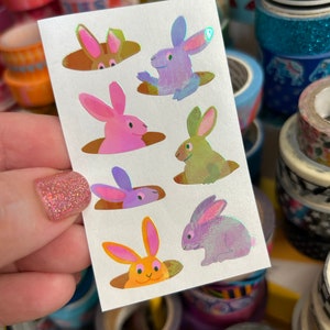 May include: A sheet of iridescent stickers featuring seven bunnies peeking out of holes. The bunnies are in various colors, including pink, purple, green, and orange.