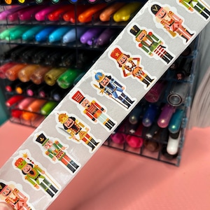 May include: A sheet of colorful nutcracker stickers with a silver background. The stickers feature a variety of nutcracker designs, including a red and gold nutcracker, a green and gold nutcracker, and a blue and gold nutcracker.