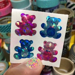 May include: A sheet of four iridescent holographic teddy bear stickers. The bears are pink, blue, blue, and pink. The bears are wearing a bow tie.