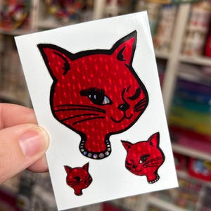 May include: A red cat sticker with a black outline and a winking eye. The cat has a dotted texture and a small necklace. Two smaller cat stickers are also visible.