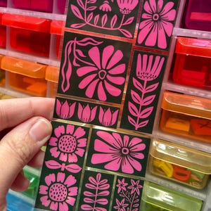 May include: A sheet of floral stickers with a variety of pink flower designs on a black background. The stickers are held in front of a colorful storage container. The text "FLORAL IMPRESSIONS" and "www.pipsticks.com" is visible.
