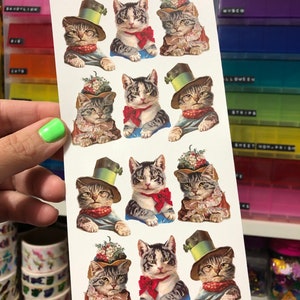 May include: A sheet of stickers featuring nine adorable cats wearing hats and accessories. The cats are depicted in a vintage style with intricate details. The stickers are perfect for adding a touch of whimsy to journals, planners, or any other creative project.