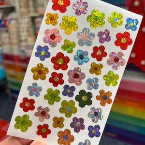May include: A sheet of colorful flower stickers. The stickers feature a variety of colors, including red, yellow, pink, blue, and green, with a holographic, iridescent effect. The flowers are in various sizes and are arranged on a white background.