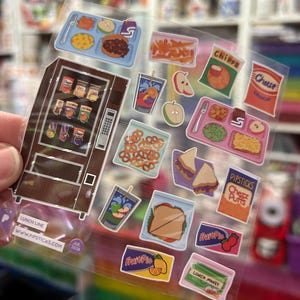 May include: A clear sticker sheet featuring a brown vending machine and various school lunch items including trays with food, sandwiches, bags of chips labeled "Chipps" and "Cheez Puffs," fruit slices, pretzels, and juice boxes. Text includes "LUNCH LINE" and "WWW.PIPSTICKS.COM".