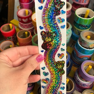 May include: A sheet of rainbow holographic stickers with a repeating pattern of a rainbow, hearts, and bears. The stickers are on a clear sheet with a white border.
