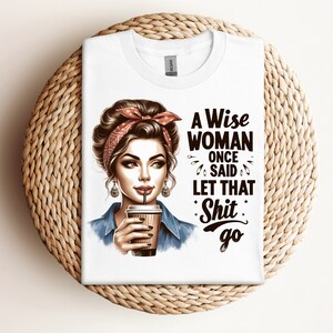 May include: White t-shirt with a graphic of a woman wearing a red bandana and holding a coffee cup. The text on the shirt says "A Wise Woman Once Said Let That Shit Go."