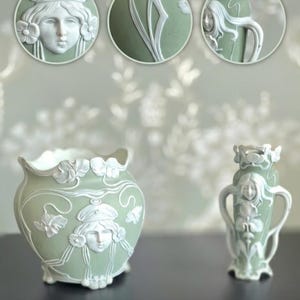 Puede incluir: Jarrones de cerámica verde salvia con diseños en relieve blancos. Los jarrones presentan rostros femeninos clásicos, detalles florales y líneas curvas. Uno es una maceta redonda, el otro es un jarrón más pequeño con asas.