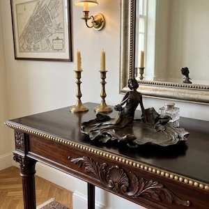 Op de afbeelding: Een donkerbruine houten console met sierlijke houtsnijwerken en gouden accenten. Een bronzen beeld van een vrouw ligt op een decoratieve schaal. Twee messing kandelaars met brandende kaarsen staan op de tafel.