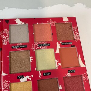 May include: A red eyeshadow palette with nine square pans of various colors, including gold, brown, and peach. Each shade is labeled with a name, such as "Snow White" and "Evil Queen". The palette has a decorative floral and heart design.