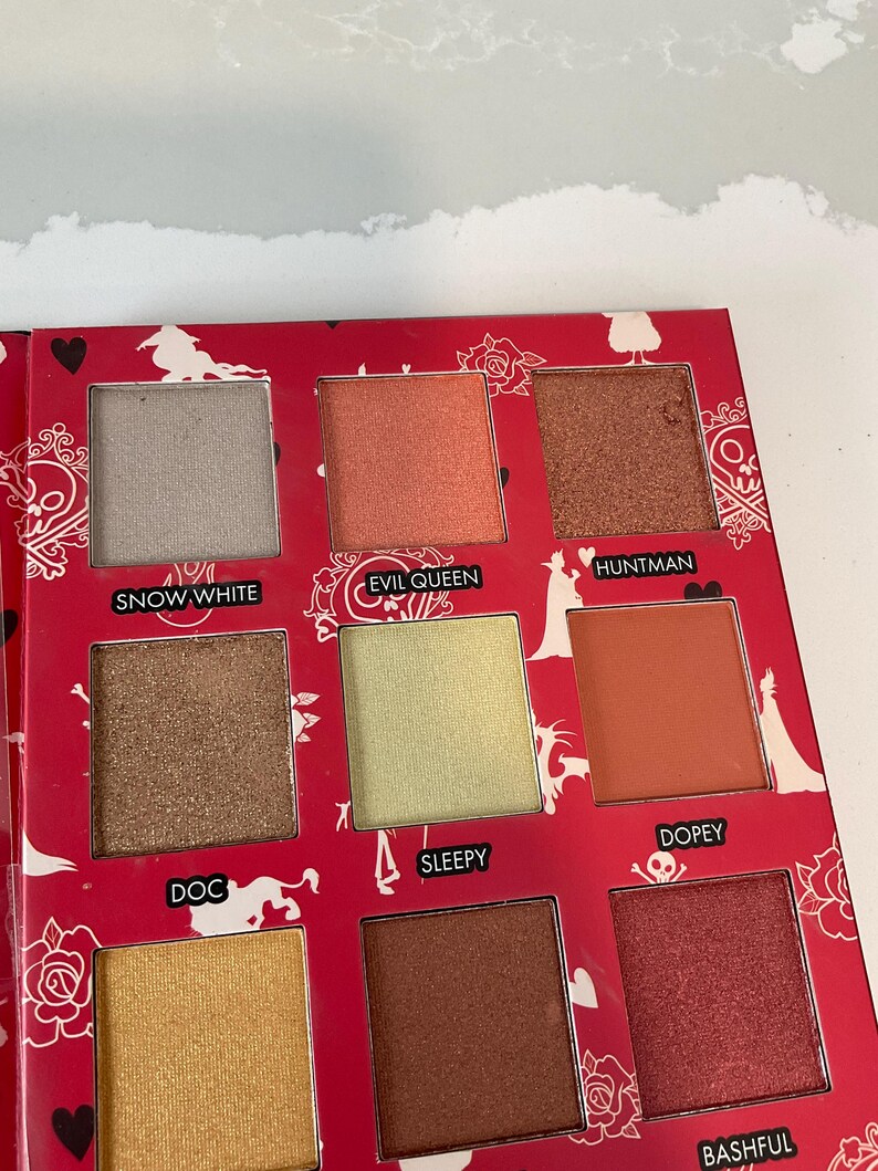 May include: A red eyeshadow palette with nine square pans of various colors, including gold, brown, and peach. Each shade is labeled with a name, such as "Snow White" and "Evil Queen". The palette has a decorative floral and heart design.