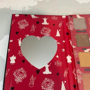 May include: Open eyeshadow palette with a red cover featuring a heart-shaped mirror. The cover is decorated with white skull and crossbones, roses, and silhouettes. The palette includes various eyeshadow colors.