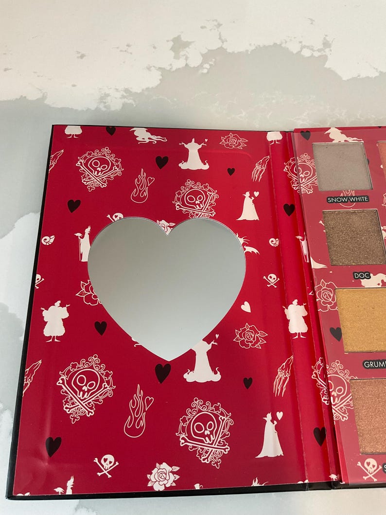 May include: Open eyeshadow palette with a red cover featuring a heart-shaped mirror. The cover is decorated with white skull and crossbones, roses, and silhouettes. The palette includes various eyeshadow colors.