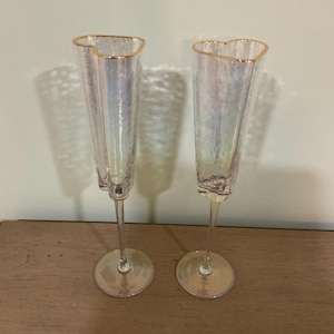 Peut inclure: Deux flûtes à champagne irisées avec des bords dorés et des ouvertures en forme de cœur. Les flûtes sont sur une surface en bois.