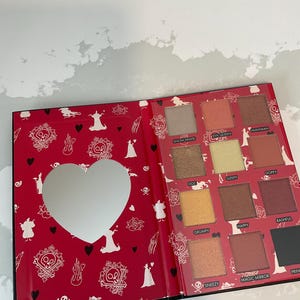 May include: Open eyeshadow palette with a red cover featuring a heart-shaped mirror and various eyeshadow colors. The palette includes shades like Snow White, Evil Queen, and Dopey. The cover has a pattern of skulls, hearts, and silhouettes.
