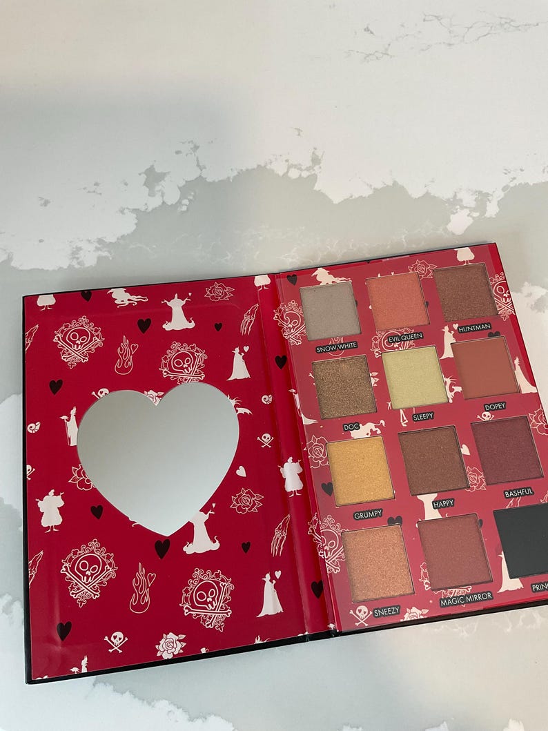 May include: Open eyeshadow palette with a red cover featuring a heart-shaped mirror and various eyeshadow colors. The palette includes shades like Snow White, Evil Queen, and Dopey. The cover has a pattern of skulls, hearts, and silhouettes.