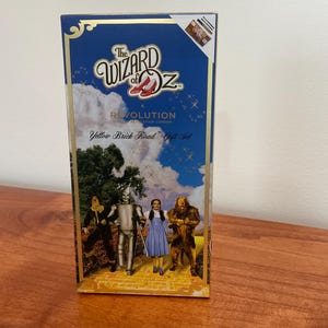 Op de afbeelding: Een blauwe en gouden doos met de woorden "The Wizard of Oz" en "Yellow Brick Road Gift Set" erop gedrukt. De doos toont een afbeelding van Dorothy, de Vogelsverschrikker, de Tin Man en de Laffe Leeuw die over de gele bakstenen weg lopen.
