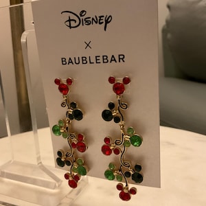 May include: A pair of gold dangle earrings featuring a Mickey Mouse head design with red, green, and black crystals. The earrings are displayed on a white card with the text "Disney x BaubleBar".