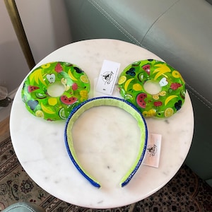 May include: A set of fruit-themed mouse ear headbands with a matching headband. The ears are green with colorful fruit slices. The headband is blue and green. A white tag with a logo is visible.