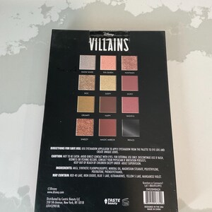 May include: A black eyeshadow palette featuring the word "VILLAINS" and Disney branding. The palette contains twelve eyeshadow shades with names like "Snow White," "Evil Queen," and "Prince." The back of the palette includes safety instructions and ingredient information.