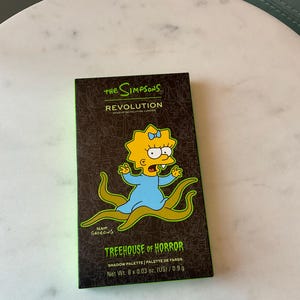 May include: A green and black makeup palette featuring a cartoon image of Lisa Simpson from The Simpsons. The palette is titled "Treehouse of Horror" and is by Revolution Makeup. The palette contains 8 eyeshadows.