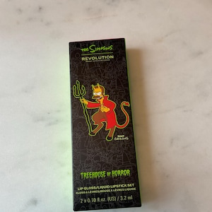 Peut inclure: Une boîte noire et verte avec une image de bande dessinée d'Homer Simpson habillé en diable. La boîte contient un ensemble de deux gloss à lèvres et rouges à lèvres liquides de la collection The Simpsons Revolution Treehouse of Horror. La boîte indique "Lip Gloss/Liquid Lipstick Set" et "2 x 0.10 fl.oz. (US)/3.2 ml".