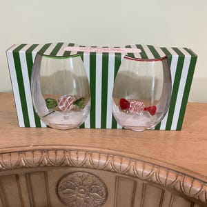May include: Two stemless wine glasses with candy decorations inside, presented in a green and white striped box. One glass has a green rim and a candy with green and white stripes. The other glass has a red rim and a red candy.