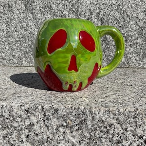 May include: A ceramic mug with a skull design. The mug is primarily green with red accents forming the eyes and mouth of the skull. The mug has a rounded shape with a handle on the side.