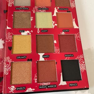 May include: A red eyeshadow palette with 12 shades, each labeled with a name. Colors range from light beige to black, including gold, orange, and brown tones. The palette has a decorative design with silhouettes and text.