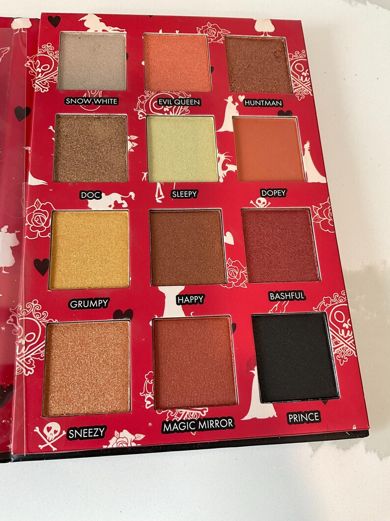 May include: A red eyeshadow palette with 12 shades, each labeled with a name. Colors range from light beige to black, including gold, orange, and brown tones. The palette has a decorative design with silhouettes and text.