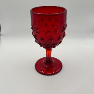 May include: A ruby red vintage goblet with a textured starburst pattern on the bowl. The stem and base are also red. The glass is likely made of pressed glass.