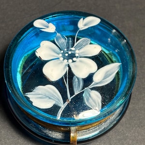May include: A round, blue glass trinket box with a hinged lid. The lid features a hand-painted white flower with delicate leaves. The box has a gold-colored metal rim and clasp, and the interior is mirrored.