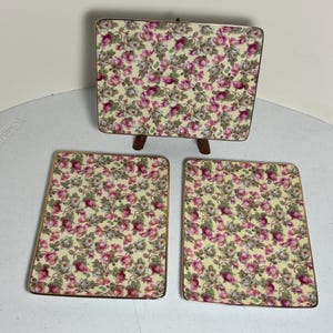 May include: A set of three decorative items with a floral pattern. The set includes two rectangular coasters and a small rectangular tray on a wooden stand. The floral pattern features pink roses and green leaves on a cream background, with gold trim.
