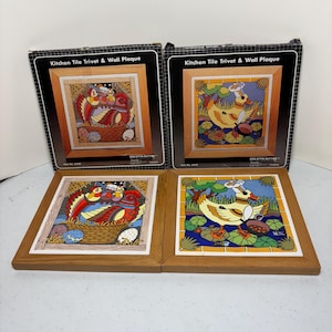 May include: Two decorative kitchen tile trivets and wall plaques. One features a colorful bird scene in a basket with eggs, the other a duck in a pond. Both are framed in wood and come in their original boxes. The boxes read "Kitchen Tile Trivet & Wall Plaque."