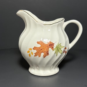 May include: White ceramic creamer with a gold rim and a fall leaf design. The leaves are orange, red, and green.