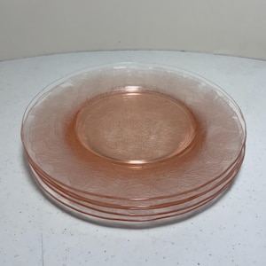May include: A stack of four pink glass plates. Each plate has a delicate floral pattern around the rim. The plates are a soft, translucent pink color, and are stacked on top of each other.