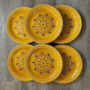 May include: Six mustard-yellow ceramic plates with a scalloped edge. Each plate features a central black floral design with radiating lines and a circular pattern. The plates are arranged in two rows of three.