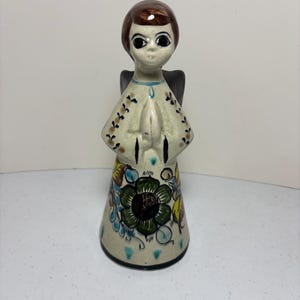May include: A ceramic angel figurine with brown hair, large eyes, and folded hands. The angel wears a cream-colored robe adorned with floral patterns in green, blue, and yellow. Black wings are visible behind the figure.