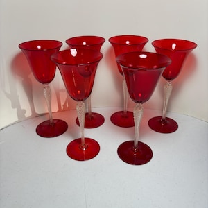 May include: Six red wine glasses with clear, twisted stems and round bases. The glasses have a tulip-shaped bowl and are arranged in a semi-circle. The glasses are made of glass and are on a white surface.