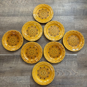 May include: A set of eight yellow ceramic plates with a black floral design in the center. The plates have a scalloped edge and are arranged on a wooden surface. The plates are a vintage style.
