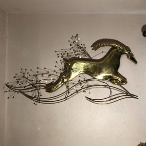 May include: A gold metal wall sculpture of an antelope leaping over stylized, wavy lines with gold dots. The antelope is in a running pose with its head turned to the side.