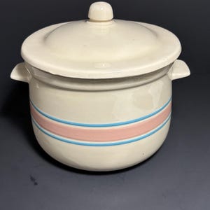 May include: Cream-colored ceramic casserole dish with a lid. The dish features a pink stripe between two blue stripes around the body. The lid has a small knob on top and the dish has two small handles.