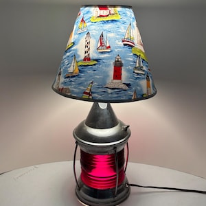 May include: A nautical-themed table lamp with a red light and a lampshade featuring sailboats and lighthouses on a blue background. The lamp has a metal base and a conical lampshade. The lamp is on a white surface.