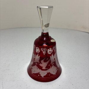 May include: A ruby red glass bell with a clear handle. The bell features etched white designs of birds and foliage. A small sticker is affixed to the handle.
