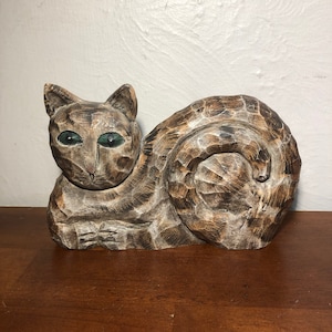 May include: A hand-carved wooden cat sculpture with a brown and gray finish. The cat is curled up with its tail wrapped around its body. The cat has green eyes and a detailed face.