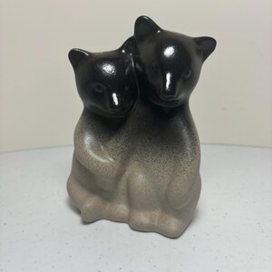May include: A ceramic figurine of two bears embracing. The bears are black on top, fading to a gray-beige at the bottom. The figurine has a glossy finish and is set against a plain background.