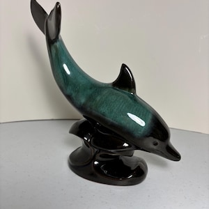 May include: A ceramic dolphin figurine in shades of teal and black. The dolphin is arched, with its tail and dorsal fin pointing upwards. The figurine is mounted on a black, wave-like base, creating a dynamic and elegant sculpture.