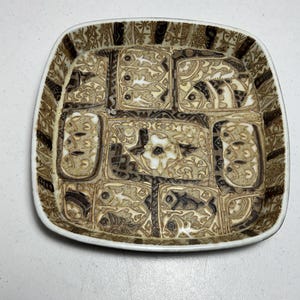 May include: A square ceramic dish with rounded corners featuring a brown and cream-colored sgraffito design. The surface is divided into rectangular panels containing stylized fish motifs, floral patterns, and geometric borders in dark brown.